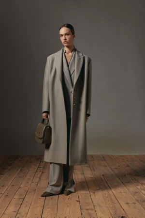 Model wearing an oversized grey wool coat holding a leather handbag against a dark backdrop