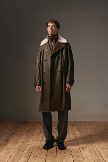 Adult male model in a full-length pose wearing a brown leather trench coat with a white fur collar and dark pants on a wood floor.