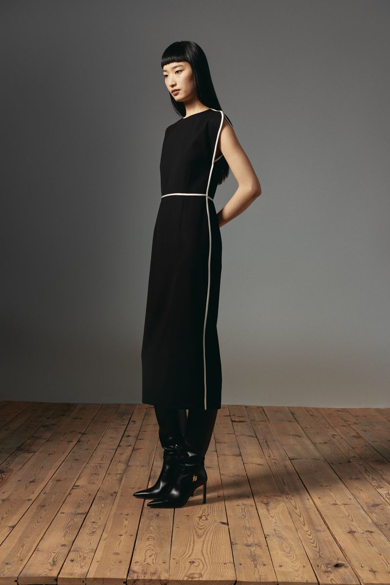 Elegant Asian woman in black midi dress with white trim, posing in black heeled boots on a wooden floor against a grey backdrop.