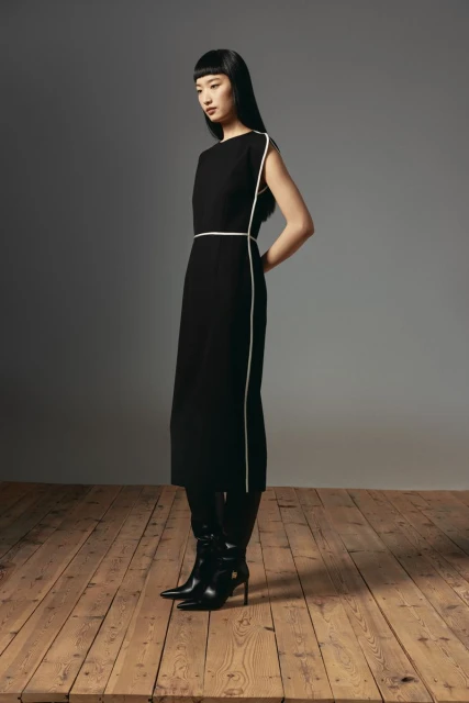 Elegant Asian woman in black midi dress with white trim, posing in black heeled boots on a wooden floor against a grey backdrop.