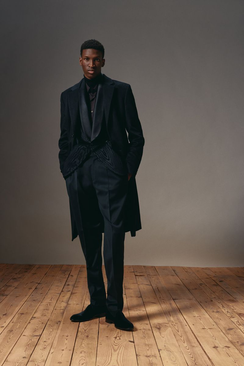 Man in dark suit poses against neutral backdrop on wood floor. He wears a tailored blazer and dress pants in a formal, elegant style.