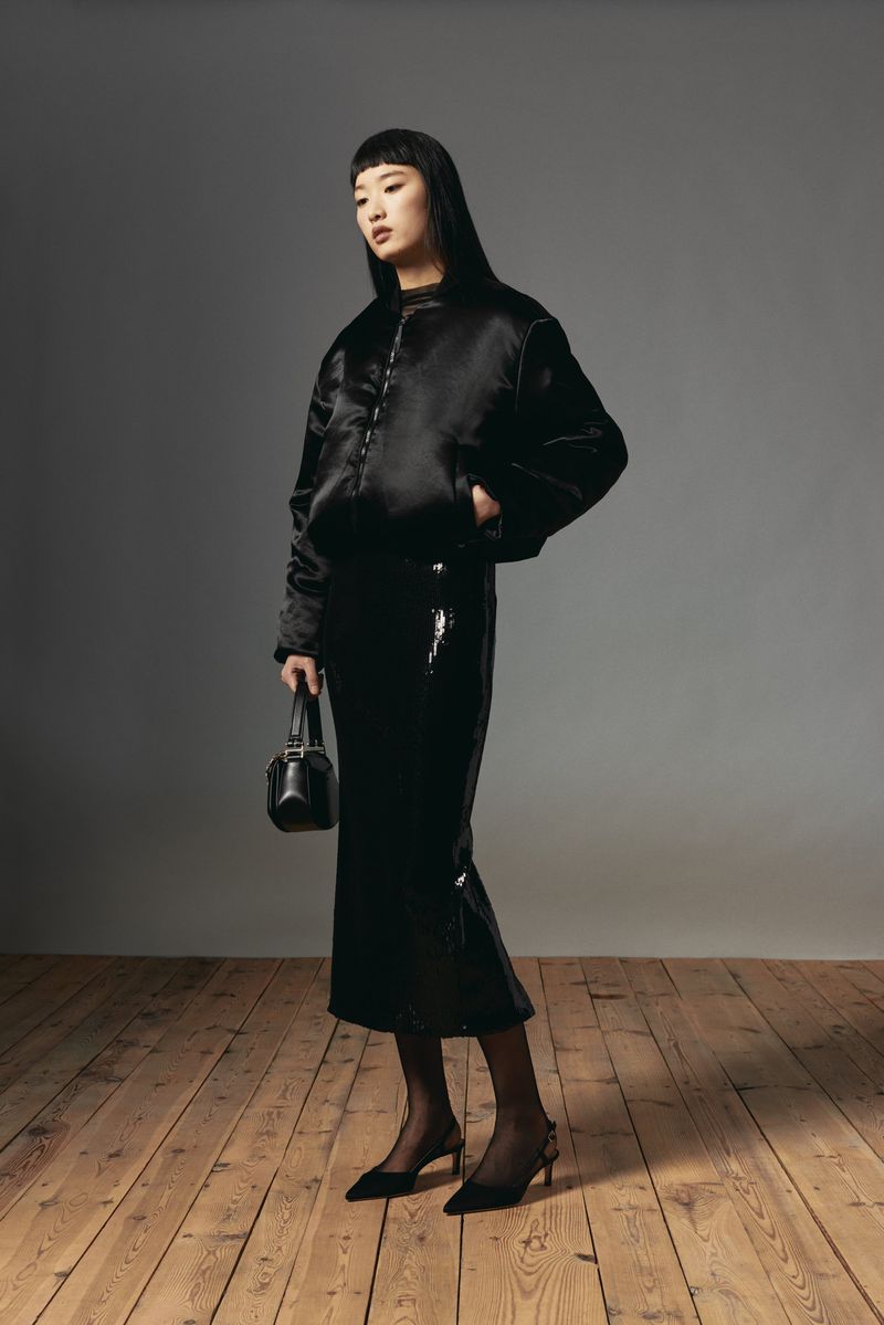 Elegant Asian woman in black jacket and skirt holding a handbag, posing against a gray background in a fashion studio shoot.