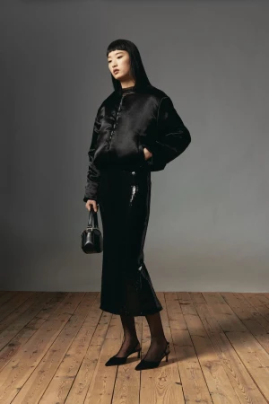 Elegant Asian woman in black jacket and skirt holding a handbag, posing against a gray background in a fashion studio shoot.