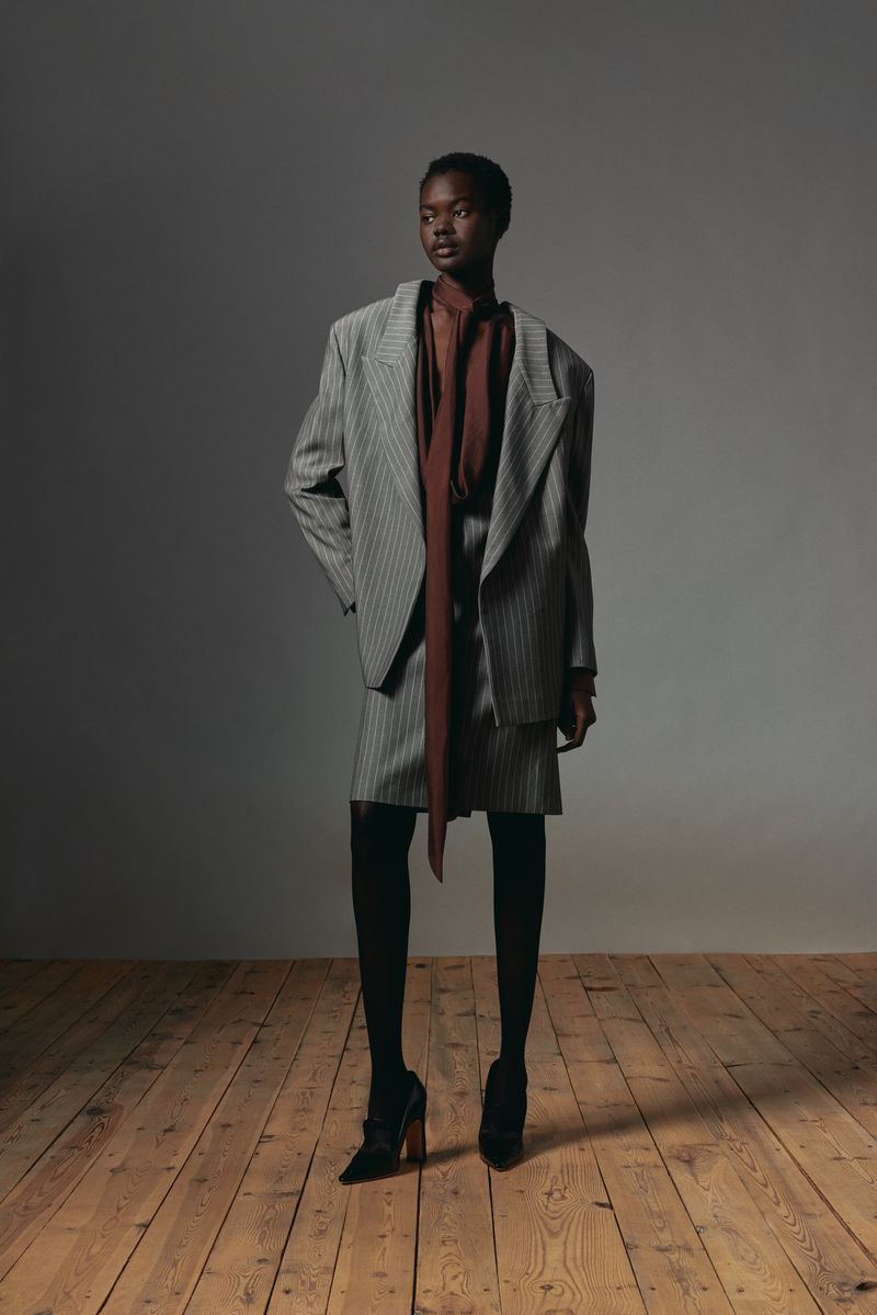 Fashion shoot of a Black woman in a grey pinstripe blazer and skirt, brown blouse, black stockings and heels, on a wood floor.
