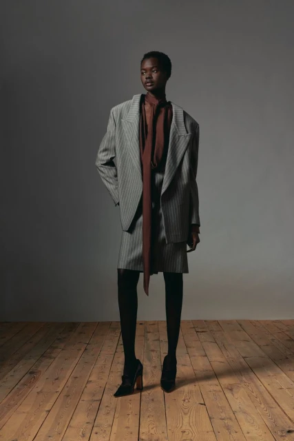 Fashion shoot of a Black woman in a grey pinstripe blazer and skirt, brown blouse, black stockings and heels, on a wood floor.