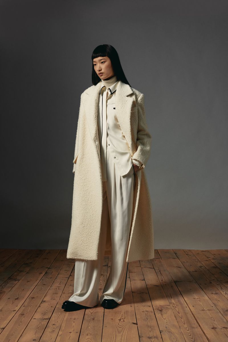Full-length fashion portrait of an Asian woman wearing an ivory coat and pants, standing on a wooden floor against a grey background.