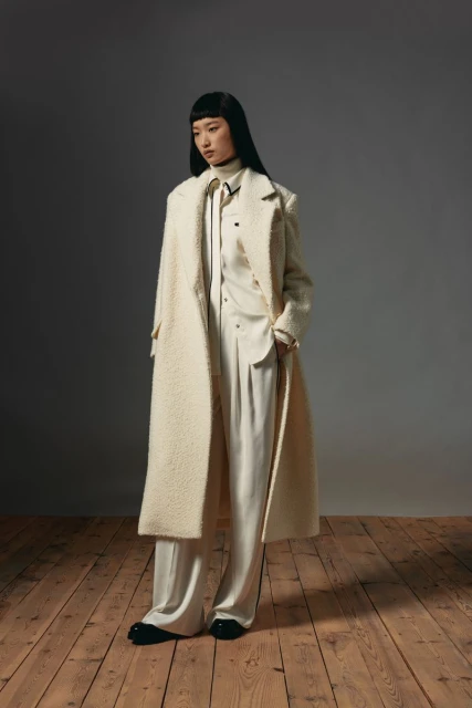 Full-length fashion portrait of an Asian woman wearing an ivory coat and pants, standing on a wooden floor against a grey background.