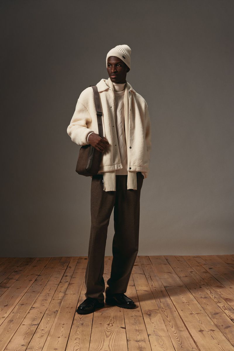 Model wearing white beanie, cream fleece jacket, and brown trousers carrying a dark crossbody bag on a wooden floor.