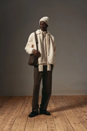 Model wearing white beanie, cream fleece jacket, and brown trousers carrying a dark crossbody bag on a wooden floor.