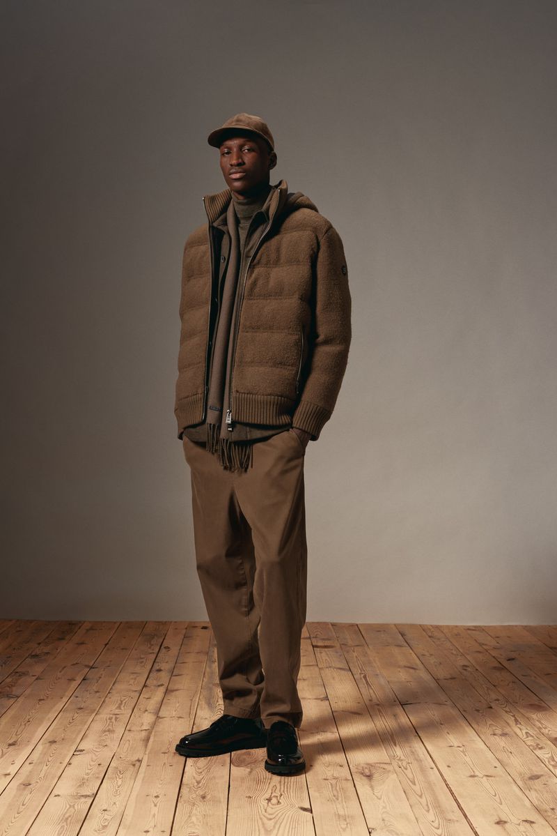 Full-length portrait of a Black man wearing a brown puffer jacket, pants, cap, and black shoes, standing on a wood floor against a grey background.
