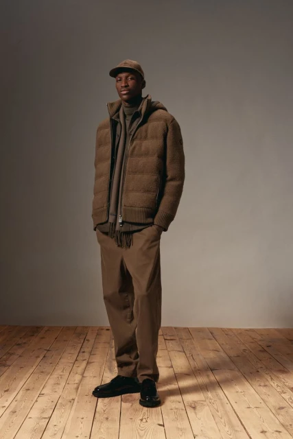 Full-length portrait of a Black man wearing a brown puffer jacket, pants, cap, and black shoes, standing on a wood floor against a grey background.