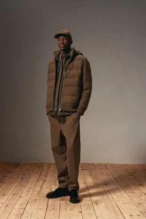 Full-length portrait of a Black man wearing a brown puffer jacket, pants, cap, and black shoes, standing on a wood floor against a grey background.