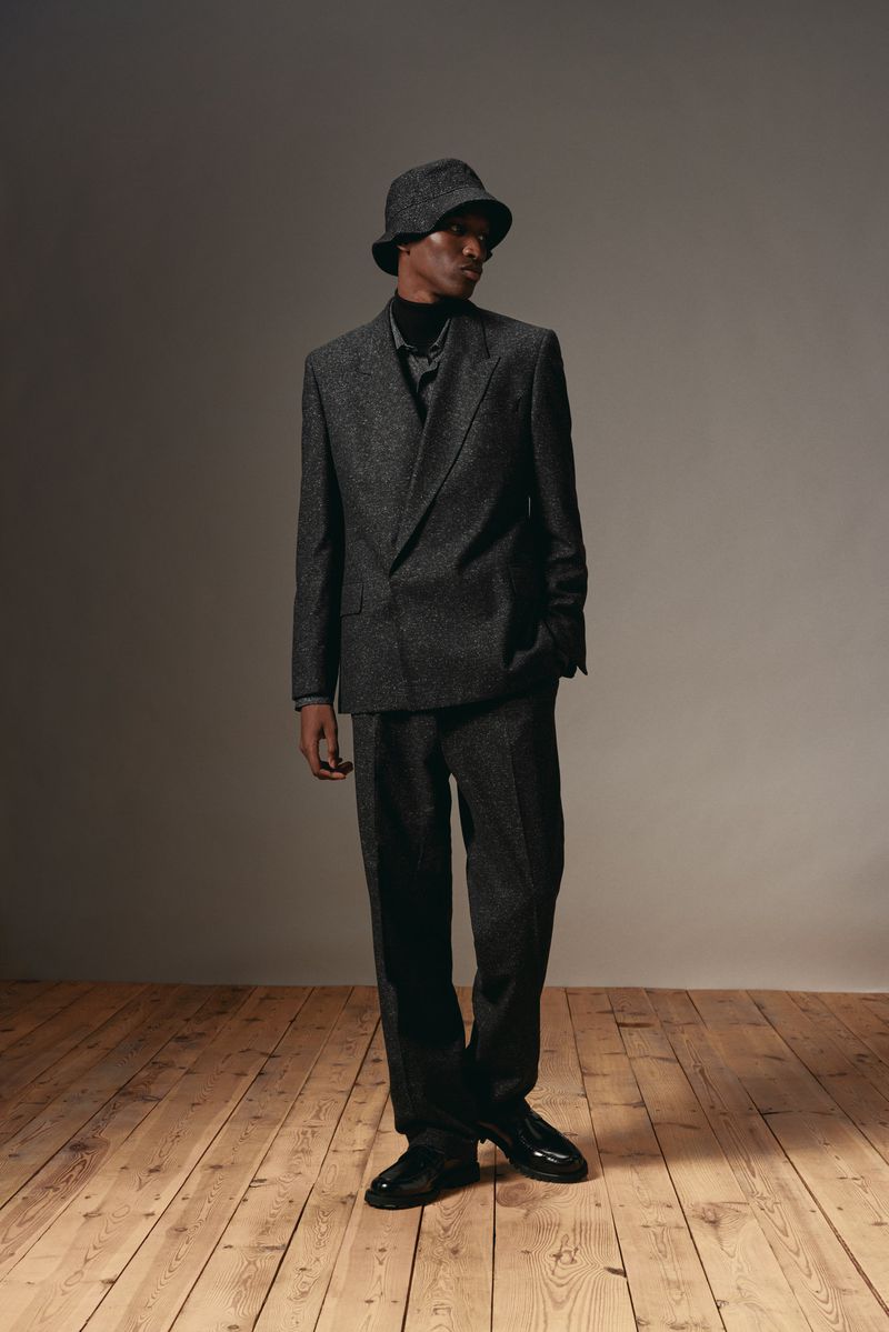 Model wearing dark grey suit and bucket hat standing on wood floor against neutral studio wall