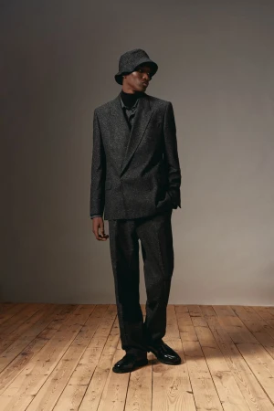 Model wearing dark grey suit and bucket hat standing on wood floor against neutral studio wall