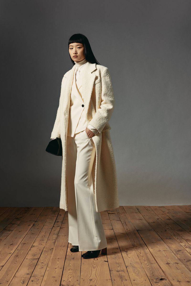 An Asian woman models a cream-colored pantsuit, turtleneck, and long coat, holding a black purse, on a wood floor with a grey background.