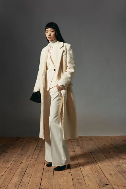 An Asian woman models a cream-colored pantsuit, turtleneck, and long coat, holding a black purse, on a wood floor with a grey background.
