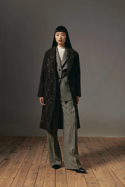 Full-length shot of an Asian woman in a striped suit and embellished coat, posing in a studio with wood floors and gray backdrop.
