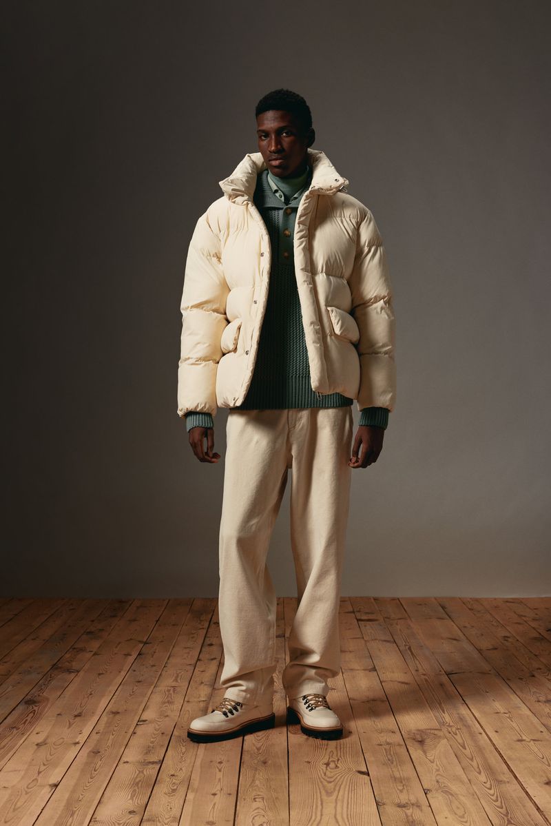 Full-length shot of a Black male model wearing a puffer jacket and cream-colored pants on a hardwood floor in a neutral indoor setting.