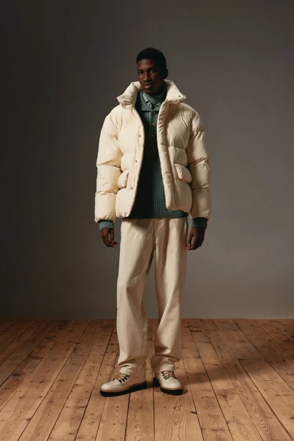 Full-length shot of a Black male model wearing a puffer jacket and cream-colored pants on a hardwood floor in a neutral indoor setting.
