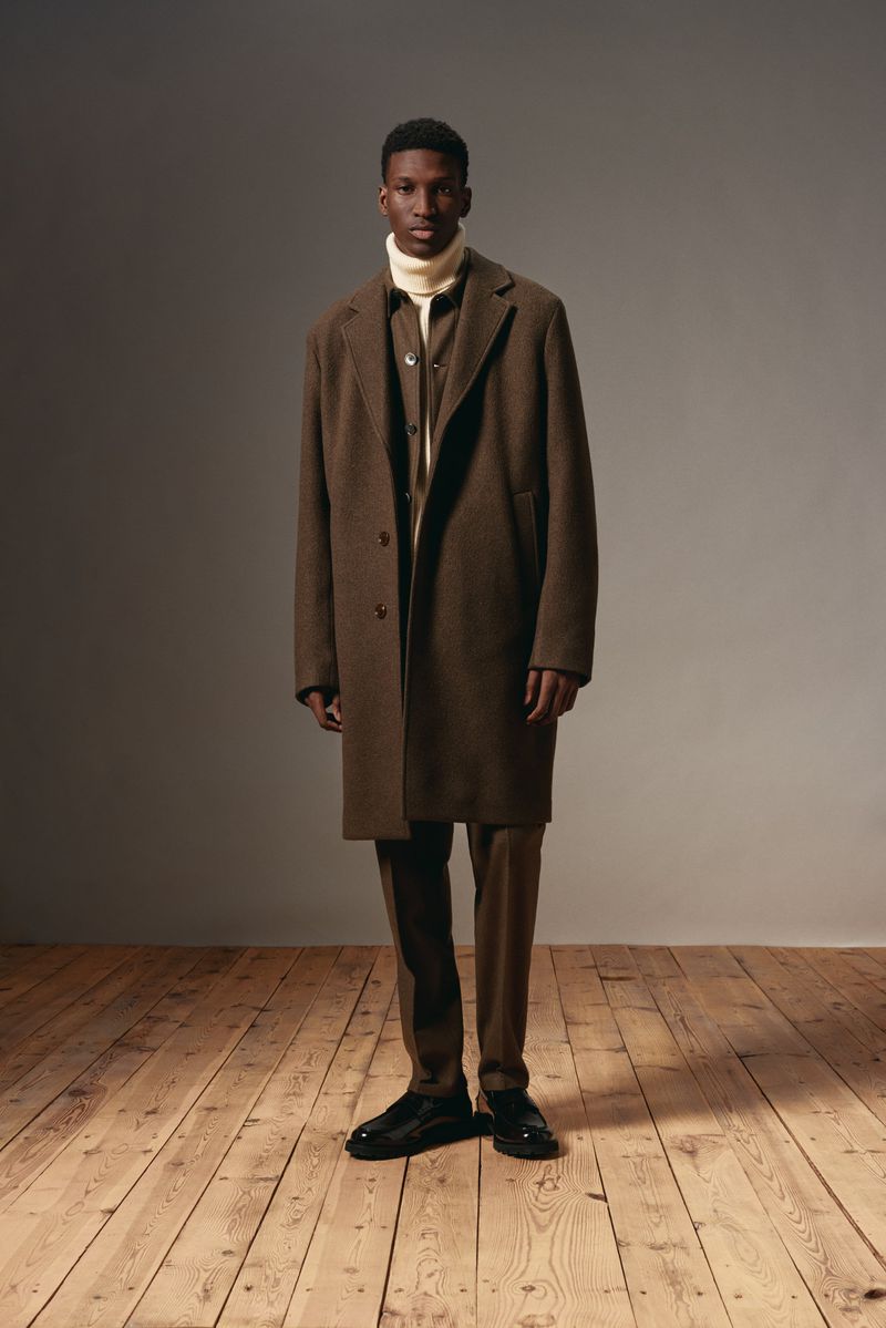 A full shot of an African American man in a brown coat and pants, posing indoors on a wooden floor against a grey background.