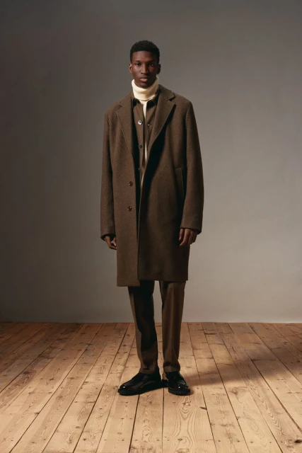 A full shot of an African American man in a brown coat and pants, posing indoors on a wooden floor against a grey background.