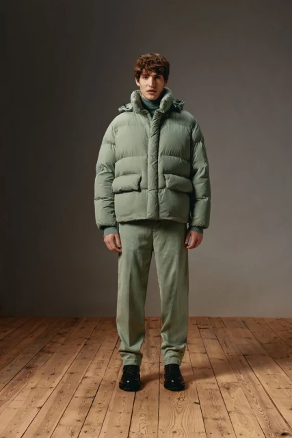 Full-length studio shot of a man in a hooded sage green puffer jacket and matching corduroy pants, standing on a wooden floor.