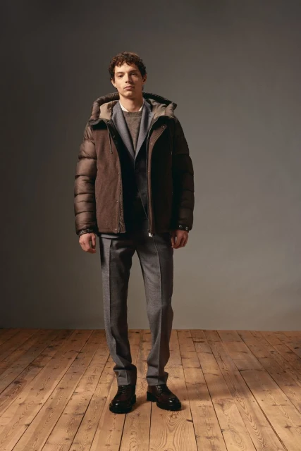 Man in gray suit and brown puffer jacket stands against gray background on wood floor in studio lighting. Fashion photography.