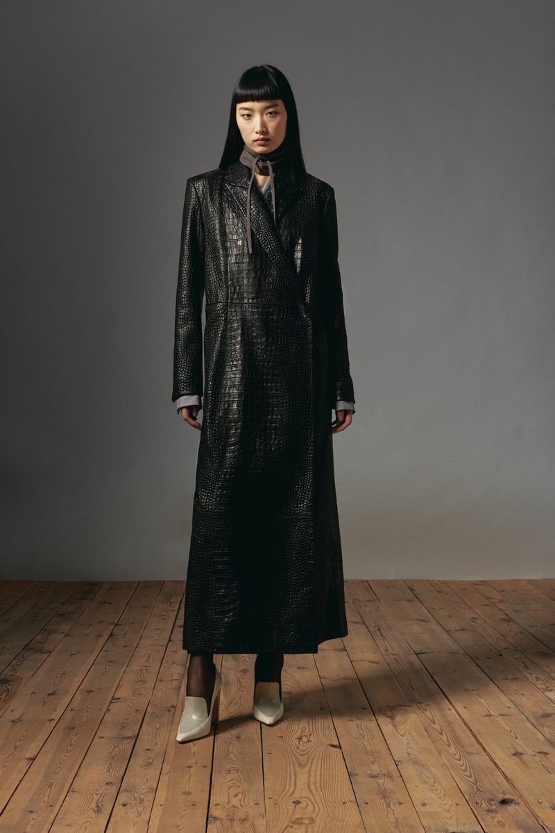 A full-length portrait of a young Asian woman in a dark, reptile-textured coat, posing in front of a gray backdrop on a wood floor. 