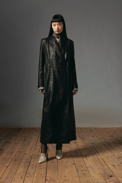 A full-length portrait of a young Asian woman in a dark, reptile-textured coat, posing in front of a gray backdrop on a wood floor. 