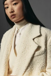 Close-up portrait of a model wearing a textured cream blazer and white turtleneck against a grey studio background