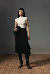 Model standing on wooden floor wearing white top and black skirt against a grey wall