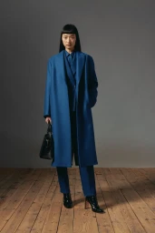 Model wearing a blue coat and holding a black handbag standing on a wooden floor against a grey wall.