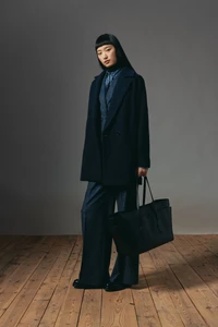 Model in dark coat and trousers holding a large black tote bag standing on a wooden floor against a grey wall.