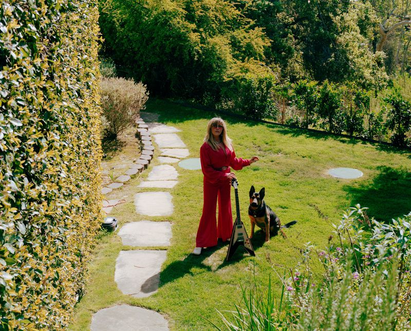 Blonde woman in a red jumpsuit stands in a garden with her German Shepherd dog and an electric guitar with a V-shaped body.