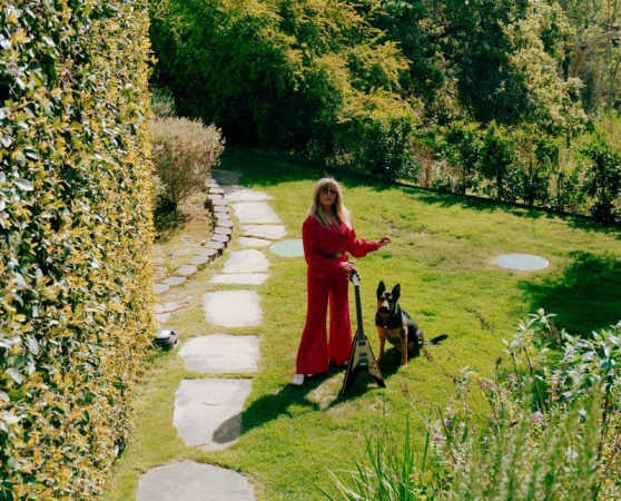 Blonde woman in a red jumpsuit stands in a garden with her German Shepherd dog and an electric guitar with a V-shaped body.