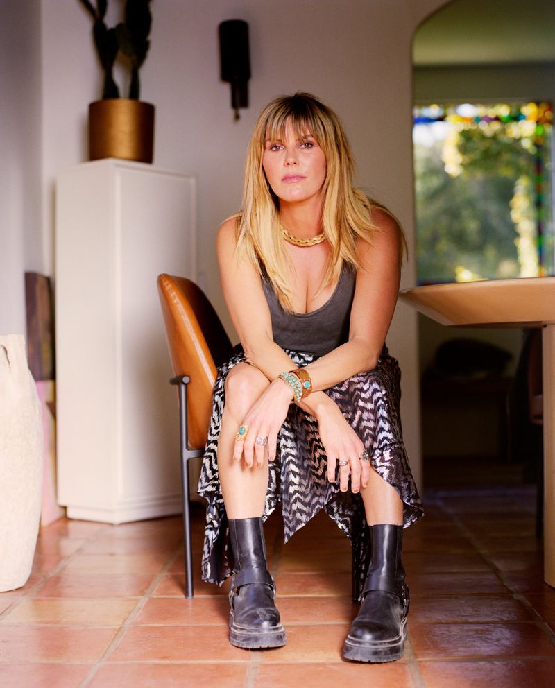 A woman with blonde hair sits in a chair, wearing a tank top, patterned skirt, jewelry, and boots, in an indoor setting.