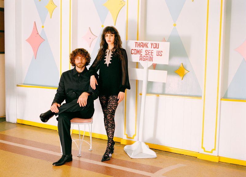 A woman and man pose indoors near a white sign; the woman wears sheer black sleeves and lace tights, while the man is seated in black.