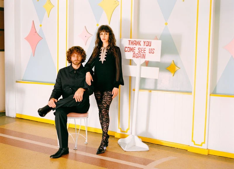 A woman and man pose indoors near a white sign; the woman wears sheer black sleeves and lace tights, while the man is seated in black.