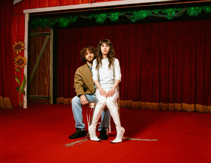 A stylish couple sits on a chair on a red stage with drawn back red curtains, posing in trendy clothes and tall white boots.