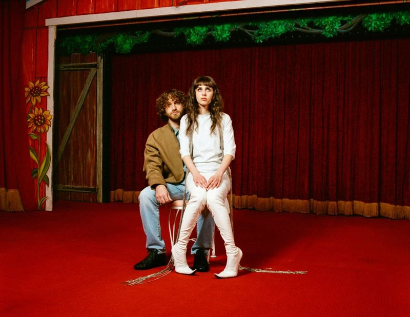 A stylish couple sits on a chair on a red stage with drawn back red curtains, posing in trendy clothes and tall white boots.