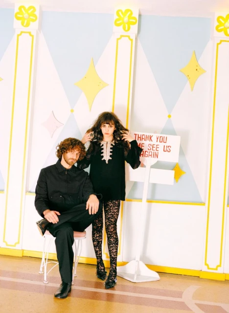 A stylish couple poses in front of a wall with diamond shapes and a hand-lettered sign, creating a vintage, fashion-forward scene.