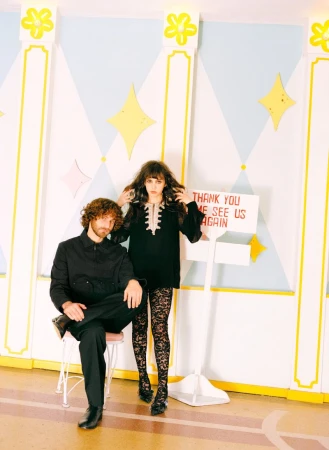 A stylish couple poses in front of a wall with diamond shapes and a hand-lettered sign, creating a vintage, fashion-forward scene.