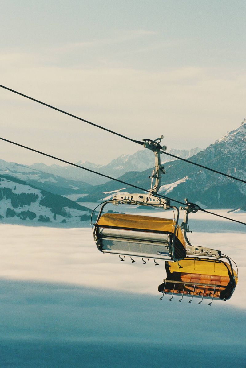 Yellow ski lift chairs ascend over fog in a mountain resort. Snowy peaks in the distance complete the scenic winter view.