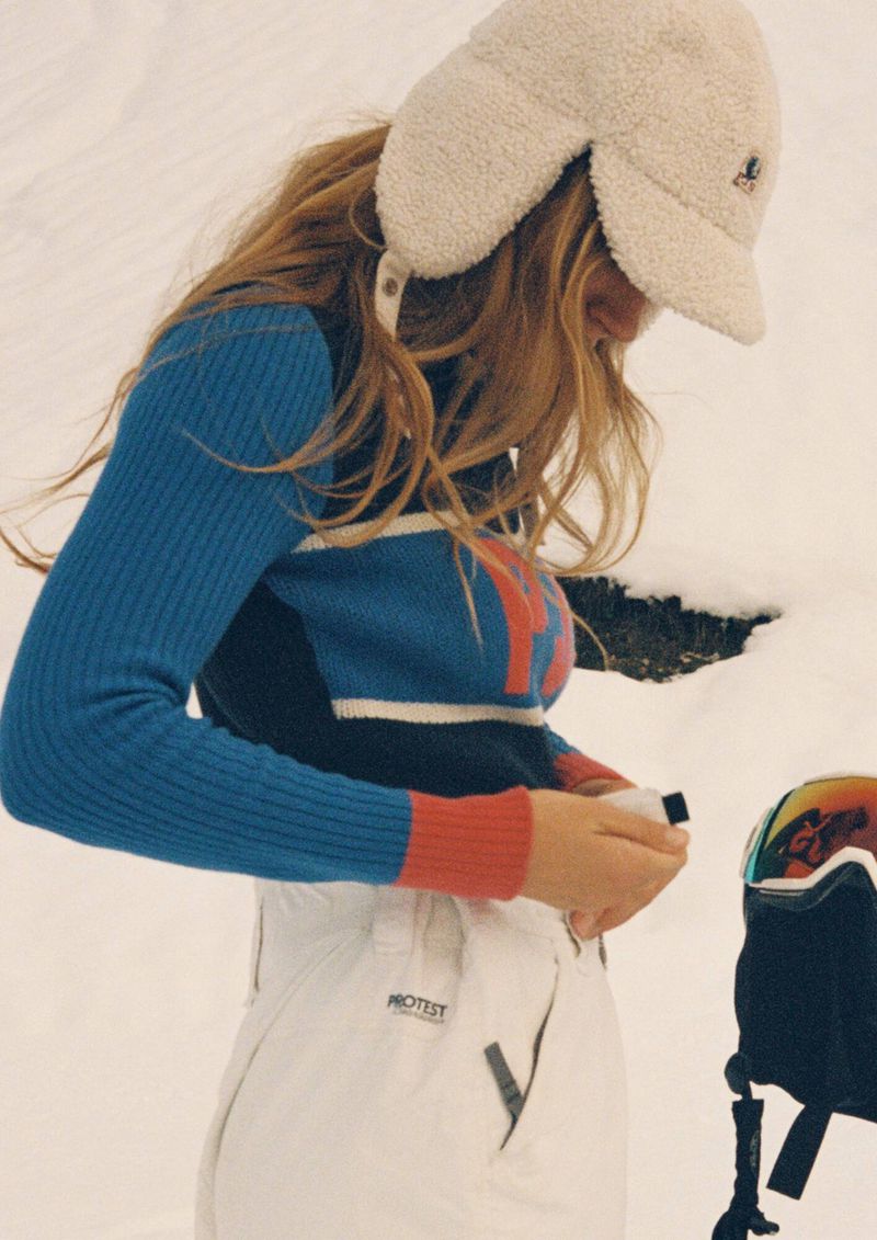 Woman in a color-blocked sweater and snow pants fastening a jacket in snowy outdoor setting with snowboard helmet visible.