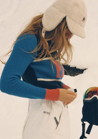 Woman in a color-blocked sweater and snow pants fastening a jacket in snowy outdoor setting with snowboard helmet visible.