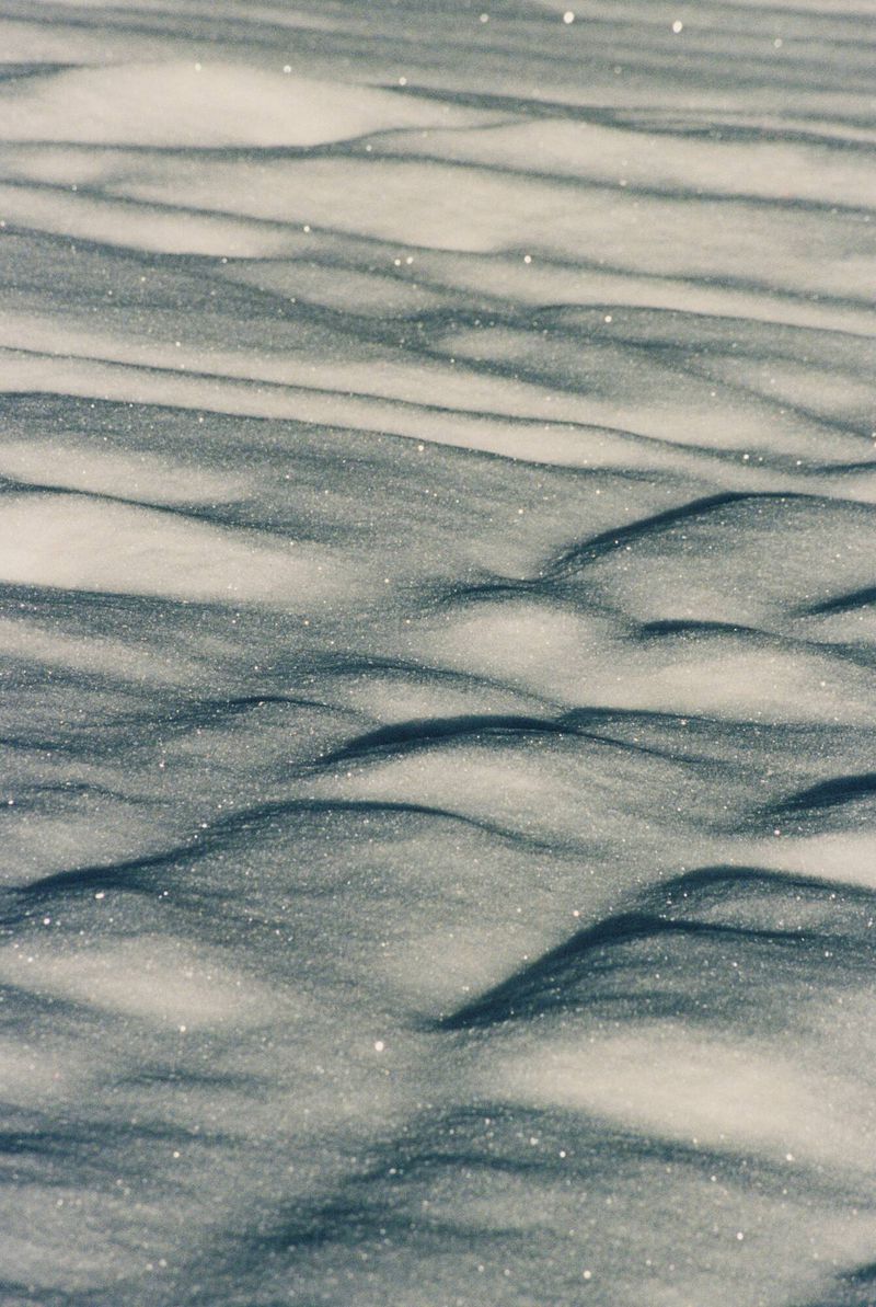 Close-up of a snow-covered surface with textured wave-like patterns created by wind. The scene is outdoors with natural lighting.