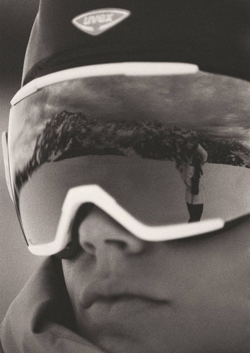 Close-up of person in ski goggles reflecting a mountain landscape and another individual, creating a monochrome, nature-focused image.