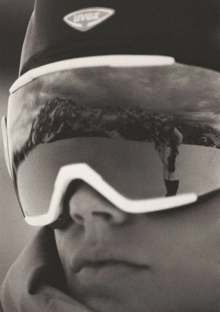 Close-up of person in ski goggles reflecting a mountain landscape and another individual, creating a monochrome, nature-focused image.