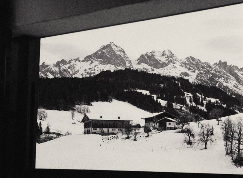 Black and white view framed through a window of alpine chalets on snow-covered slopes before dramatic rocky mountain peaks.