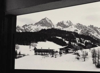 Black and white view framed through a window of alpine chalets on snow-covered slopes before dramatic rocky mountain peaks.
